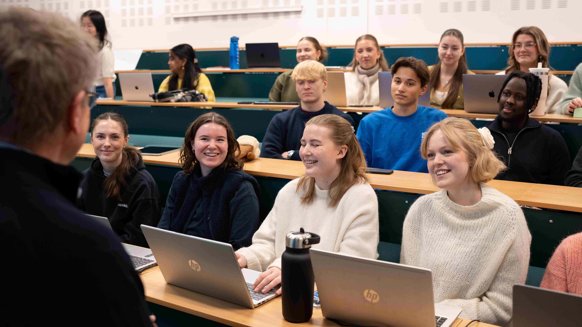 Studenter sitter på rekke og rad  i en forelesningssal, vi ser ryggen på underviseren i til venstre i bildet.