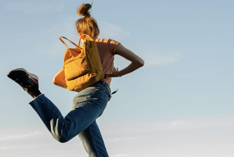 Girl with orange backpack jumping. Behind her is blue sky.