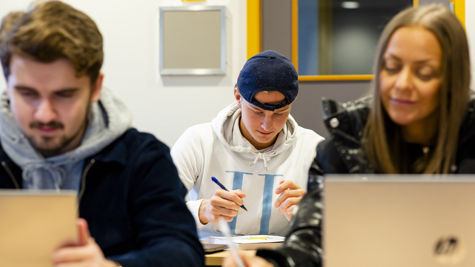Studenter i et klassrom. En mann i fokus, han har en blå caps bakfrem, og sitter med en penn i hånden. To andre i front sitter å ser på hver sin laptop, den ene en dame, den andere en mann.