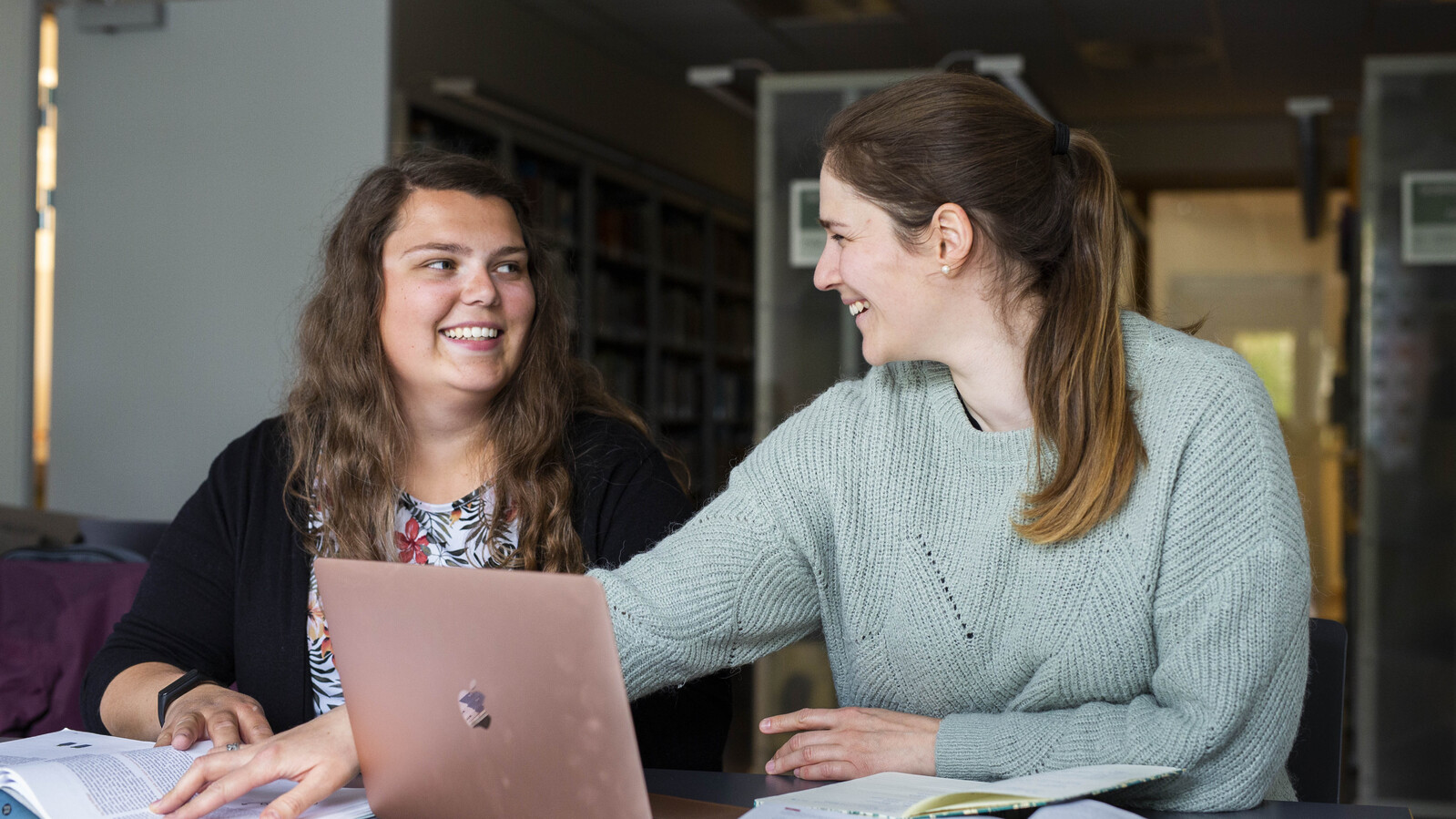 To kvinnelige studenter sitter å jobber sammen med pensum. Den ene har laptopen oppe mens og peker på noe i boken. De ser på hverandre og smiler.