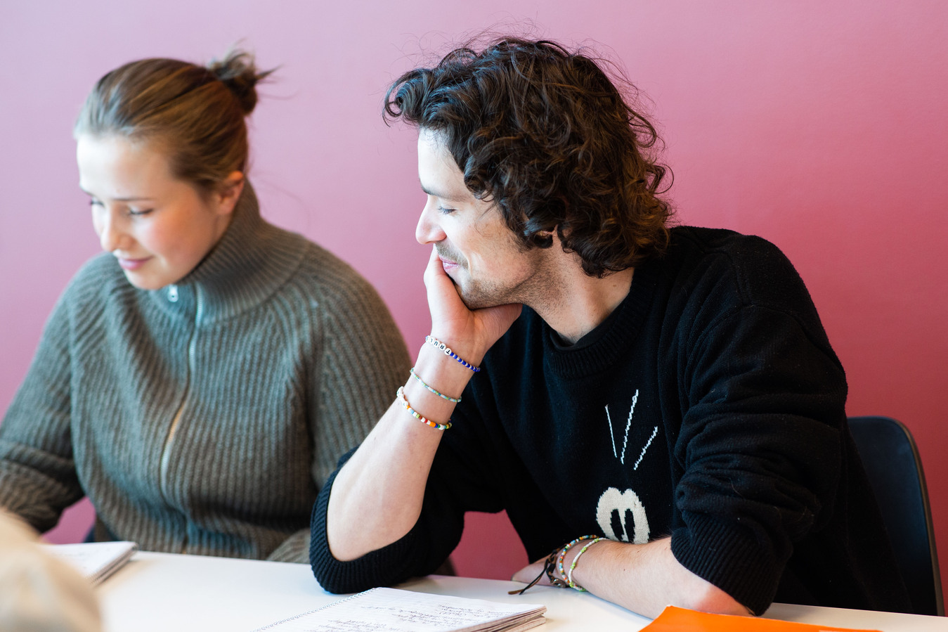 To studenter, en mann og en kvinne sitter å jobber med skoleoppgave. Veggen bak de er rød. Kvinnen har på seg en olivengrønn strikket genser med glidelås og oppsatt hår i hestehale. Mannen har på seg en svart genser som han har krøllet opp langs armene. Mannen har mørkt krøllete hår.