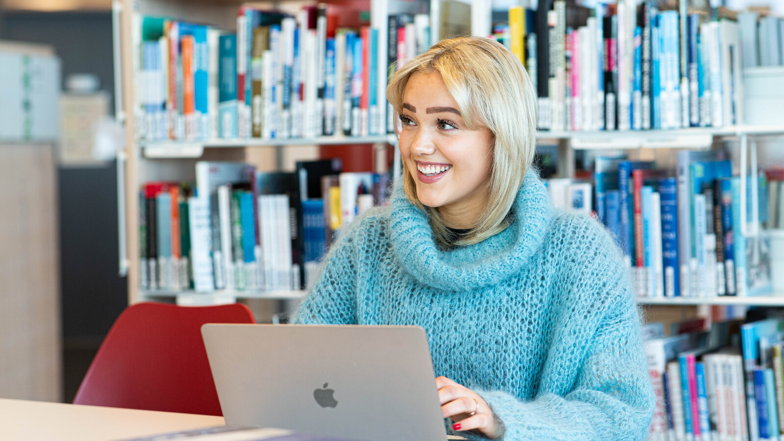 Smilende kvinne studerer på biblioteket, med en macbook fremfor seg, mange bøker i bakgrunn. Har blondt hår og en strikket genser med krage i turkis.