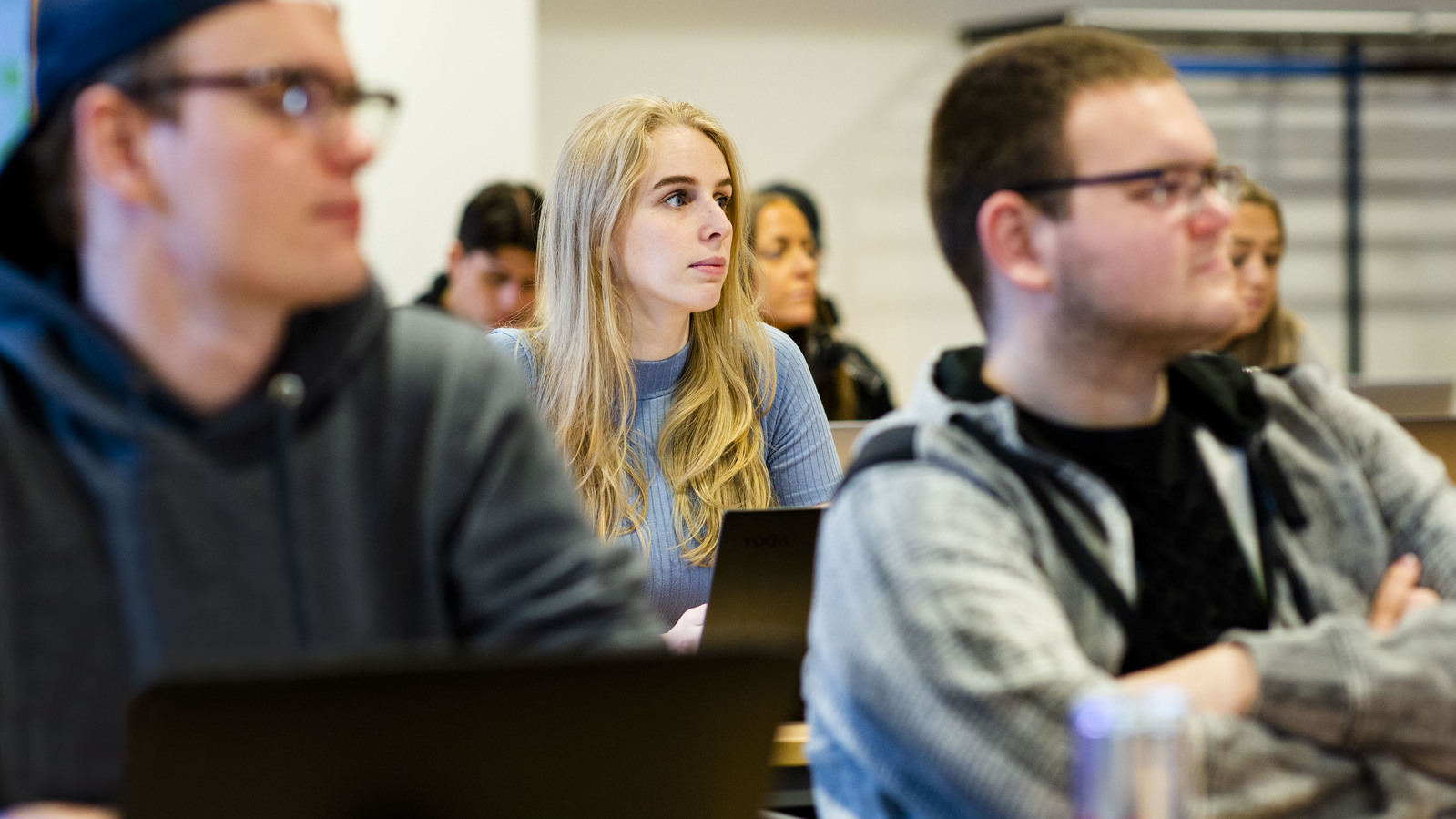 Fokus på en kvinnelig student i midten, hun har langt blondt hår og blå genser. Foran henne sitter to menn, begge har briller og ser konsentrert ut.