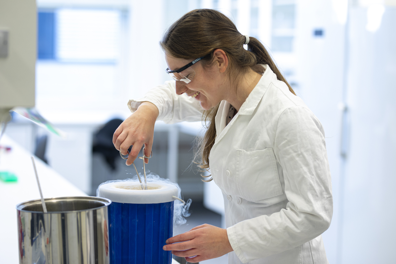 Kvinnelig student i et laboratorium med hvit frakk og vernebriller på seg, står og ser ned i en blå bøtte med noe i hånden.