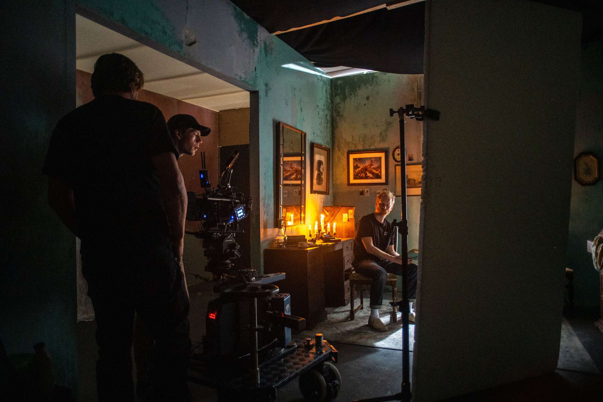 Fra en scene i forbindelse med filmproduksjon. Et hverdagsrom med en dame i en sofa og et lite bord fremfor seg, gul teppe på gulvet. En mann står i ene hjørnet foran en lenestol, han har genser og shorts på seg. I et annet hjørne står produksjonsteamet med filmkameraer, lys, microfon på pinne. Rommet har hvite dører og gråblå vegger.