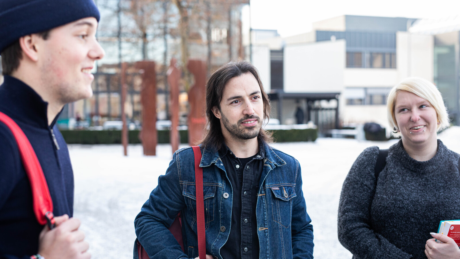 Studenter står ute, det er litt snø på bakken. To menn og en dame. En av mennen har jeansjakke på seg, den andre lue. Damen har en veske over skuldra og har blondt hår.