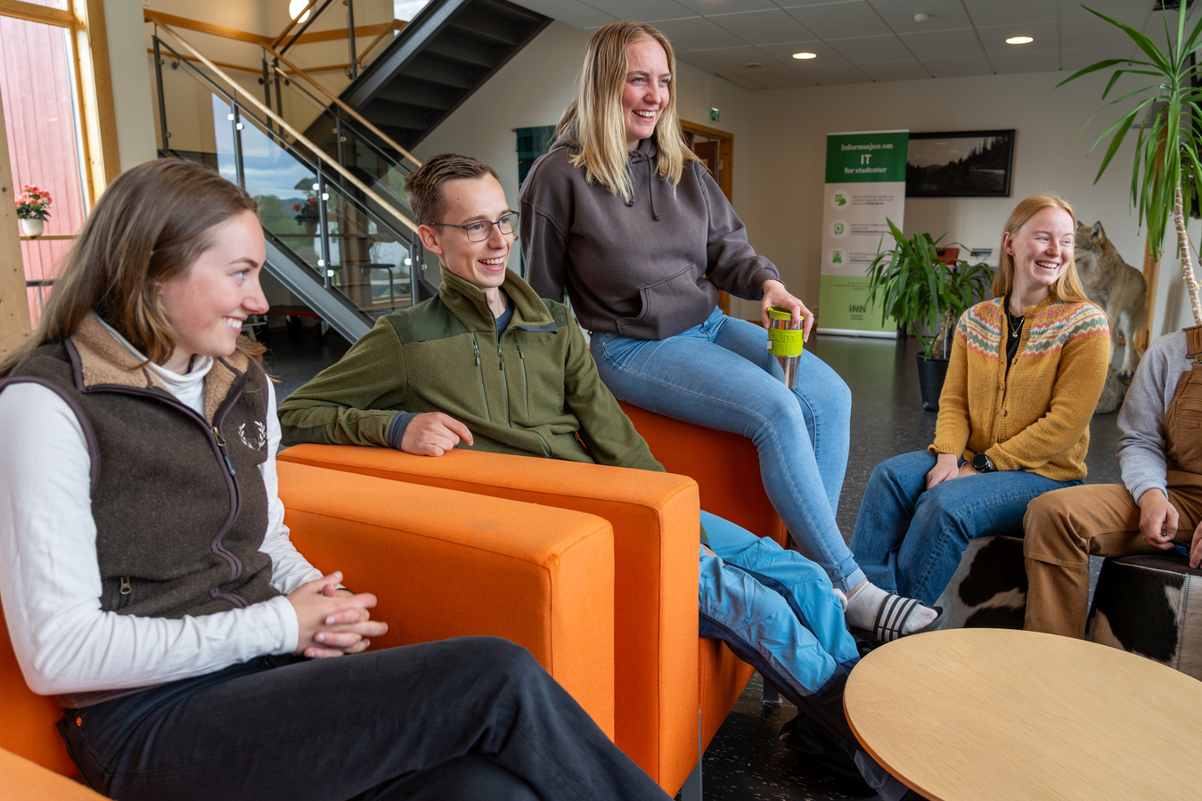 Studenter som sitter i oransje stoler. En sitter oppe på det ene armlenet. De smiler og har en samtale.