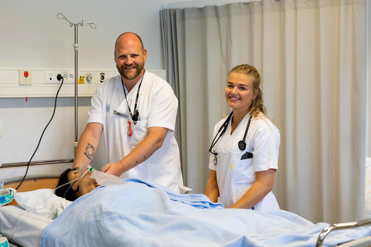 En mannlig og en kvinnelig sykepleier smiler til kamera i ført hvite klær og med stetoskop rundt halsene. Pleier en øvingsdukke i en seng på øvingslab. Blå filt over dukken.