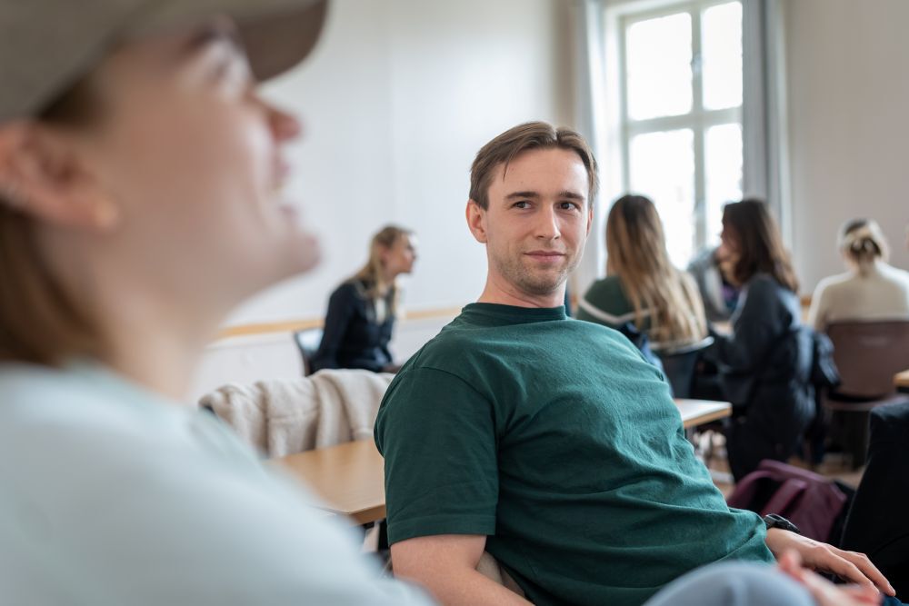 Mannlig student i grønn t-shirt sitter og ser på medstudent. Flere studenter som jobber i bakgrunn.