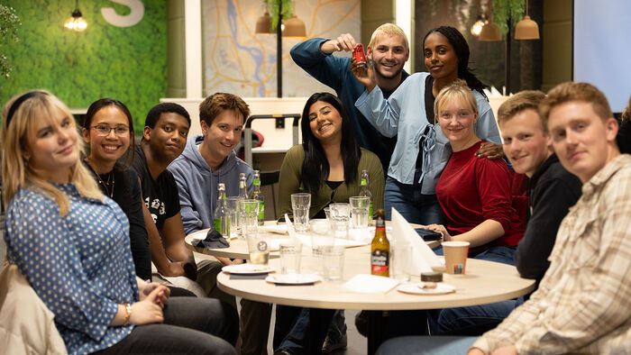 En større gruppe med glade studenter sitter rundt et bord i kantina og ser inviterende inn i kameraet.