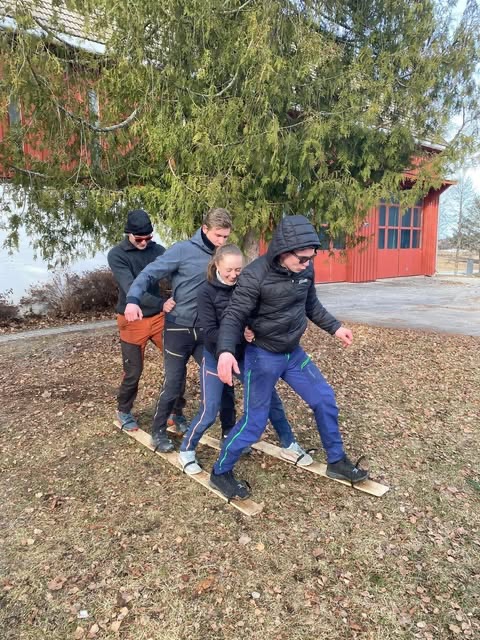 En gruppe mennesker som sitter rundt et bord, det er poser med mat og drikke på bordet. I bakgrunnen er det en rød og hvit låve