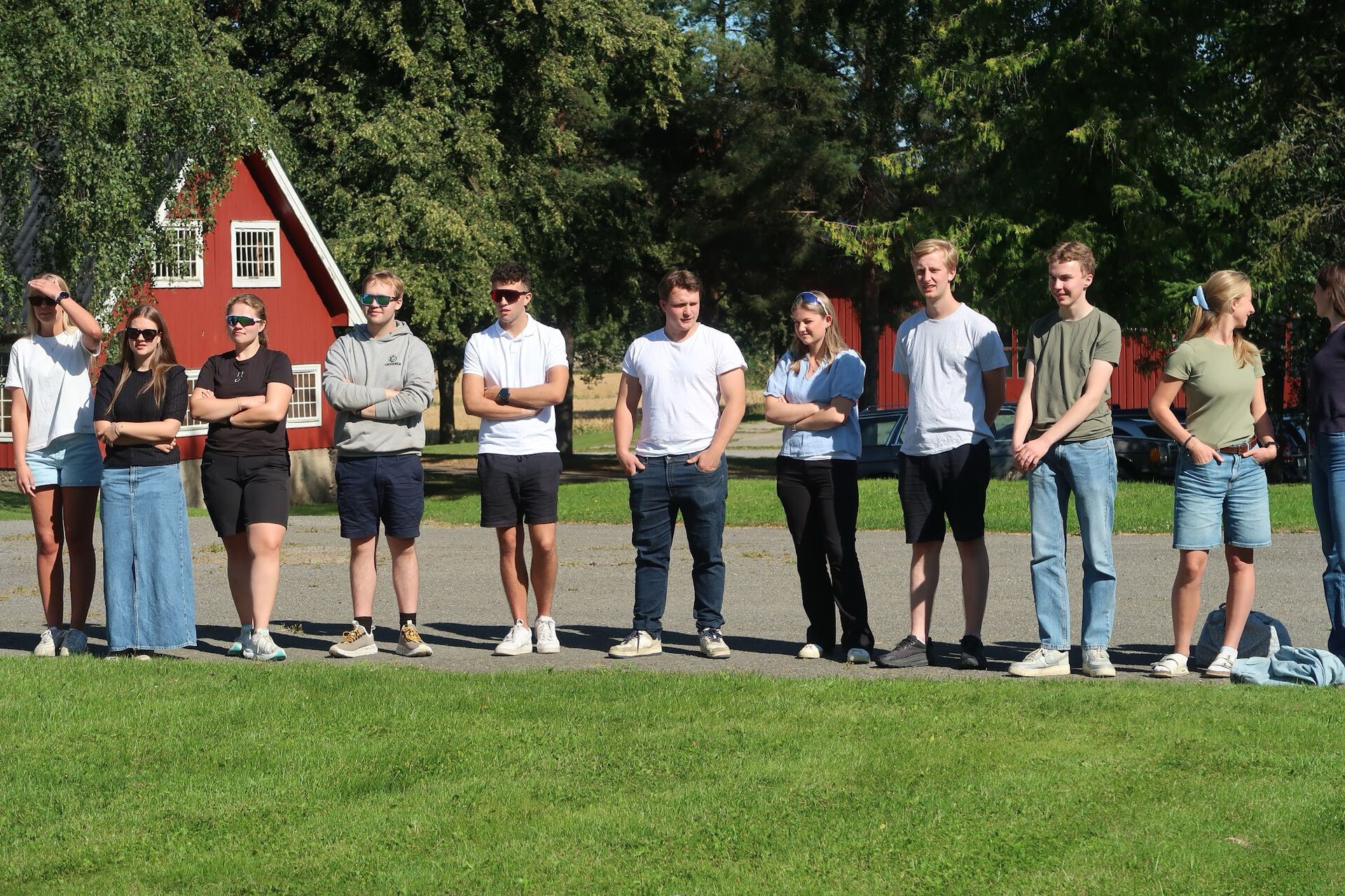 Mennesker som står på rekke ute på grønt gress. Det er sommer så de har på seg sommer klær. I bakgrunnen er det busker med grønt løv og et rødt bygg