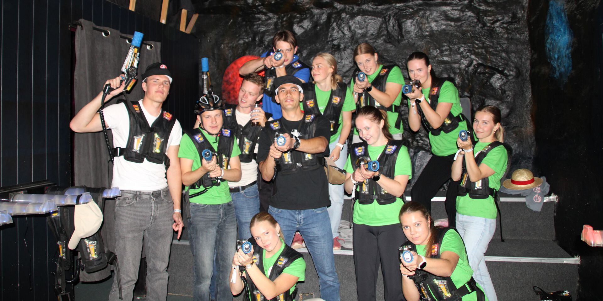 En gruppe studenter kledd i laser tag-utstyr poserer sammen etter et spill under fadderuka. Flere peker mot kamera med lasergeværene og smiler, noe som viser lek og lagånd.