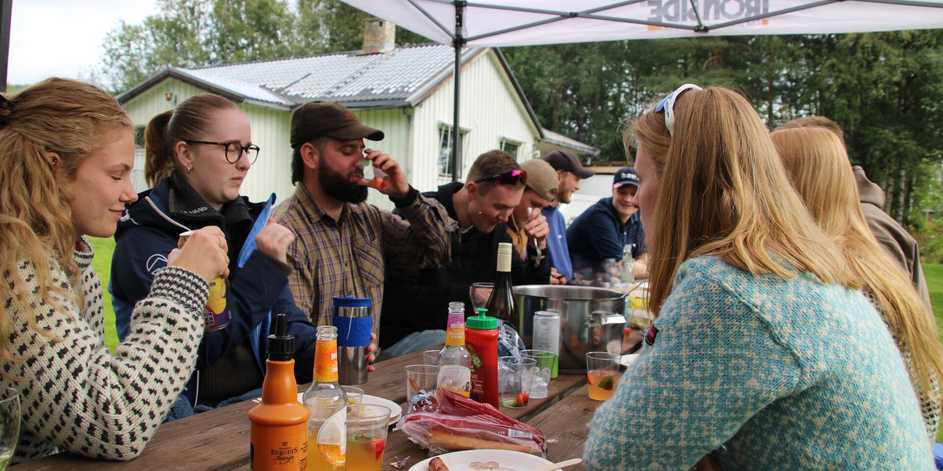 En gruppe studenter sitter ute rundt et bord under fadderuka, med mat og drikke foran seg. De prater, spiser og koser seg i uformell stemning.